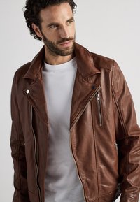 Brun lädermotorcykeljacka med dragkedja, stor krage och sidofickor med dragkedja. Texturerad yta med distinkta sömnadsdetaljer.