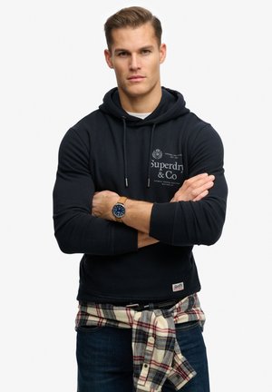 Donkerblauw sweatshirt met capuchon en een logo op de borst, gecombineerd met een geruit overhemd gestrikt om de taille en een donkere spijkerbroek.