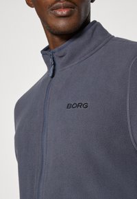 Pelēks flīsa žakets ar augstu apkakli, priekšējo rāvējslēdzēju un izšūtu "BORG" logotipu. Strukturēta auduma un kontrastējoša rāvējslēdzēja detaļas.