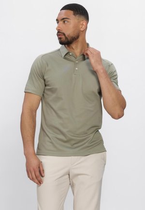 Mann mit kurzem Haar und Bart, der ein olivgrünes kurzärmeliges Poloshirt und beige Hose trägt, richtet den Kragen und blickt nach links.