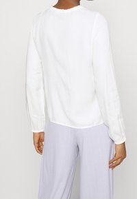 Blusa bianca a maniche lunghe con scollatura a volant e polsini elasticizzati; realizzata in un tessuto morbido, progettata per cadere leggermente sotto la vita.