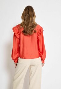 Blusa rossa a volant con maniche lunghe, in tessuto leggero e dalla consistenza morbida, abbinata a pantaloni larghi color crema. Vista posteriore mostrata.