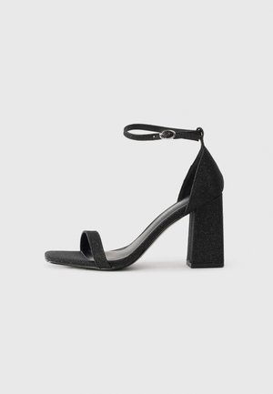 Svart högklackad sandal med glittrande yta, utrustad med ett enda tåband, ankelrem och en fyrkantig blockklack.