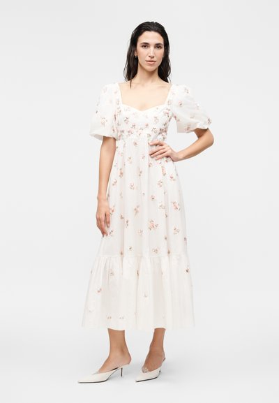 Γυναίκα στέκεται φορώντας ένα λευκό μίντι φόρεμα με φουσκωτά μανίκια και μικρά ροζ λουλουδάτα σχέδια, σε συνδυασμό με λευκές μυτερές γόβες.