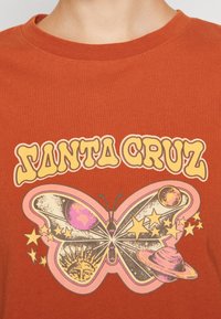 Rostfärgad bomulls t-shirt med en grafisk design av en fjäril tillsammans med planeter, stjärnor och texten "SANTA CRUZ" i retrostil.