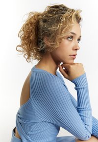 Der blaue, gerippte Pullover verfügt über einen Ausschnitt auf der Rückseite und schmal geschnittene, lange Ärmel. Der strukturierte Stoff verleiht dem Pullover durch sanfte Linien visuelles Interesse.