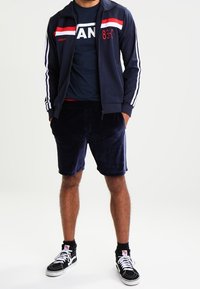 Granatowa kurtka dresowa na zamek z czerwonymi i białymi paskami, granatowa koszulka z napisem "AN" oraz granatowe szorty z weluru. Czarne sneakersy z białymi sznurowadłami.