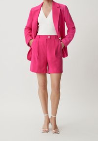 Donna in un blazer su misura rosa brillante e pantaloni corti a vita alta abbinati, con una maglietta bianca a scollo a V e tacchi a strappo bianchi.