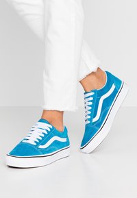 Modré semišové tenisky s bílými tkaničkami, s bílým bočním proužkem a bílou gumovou podrážkou s černým detailem. Viditelné odřené bílé džíny.