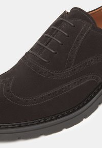 Scarpa in suede marrone scuro con design brogue, dettagli perforati, chiusura con lacci e suola in gomma a contrasto.