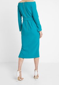 Tyrkysové midi šaty s odhalenými rameny, dlouhými rukávy, páskem v oblasti pasu a zipem na zadní straně. Nosené s bílými lodičkami na kotníkovém pásku.