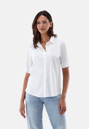 Camicia bianca a maniche corte con colletto, realizzata in tessuto morbido, con vestibilità comoda e leggero lucido. Indossata con jeans blu chiaro.