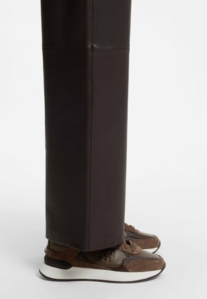 Brune læder- og ruskindsneakers med tykke hvide såler båret med mørkebrune bukser med lige ben på hvid baggrund.