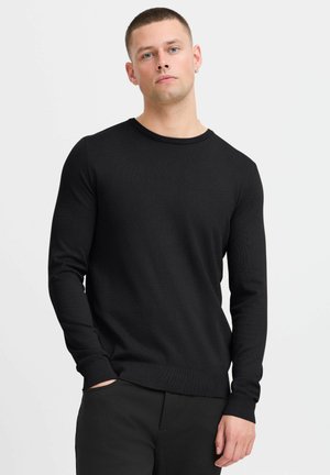 Schwarzer gerippter Pullover mit langen Ärmeln, Rundhalsausschnitt und taillierter Saum. Über einer schwarzen Hose getragen. Glatte Textur, minimalistisches Design.
