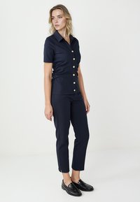 Marineblauw tweedelig ensemble met een kort mouwig hemd met gouden knopen en nauwsluitende broeken met enkelopeningen, allemaal van een gladde stof.