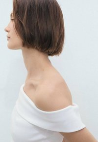 Witte off-shoulder kleding, gladde textuur, getailleerd ontwerp. Korte bruine haar, kijkt naar links, tegen een lichte achtergrond.