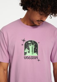 Paarse katoenen t-shirt met een zwart en groene grafische ontwerping, inclusief sterren en een gebouwsilhouet met de tekst "VOLCOM."