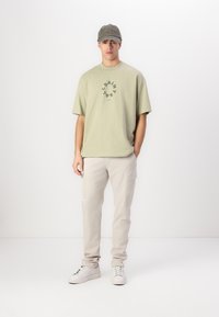 Lys oliven grønn oversized t-skjorte med sirkulært grafisk motiv, kombinert med beige cargo bukser og hvite joggesko. Caps i matchende olivenfarge.