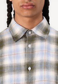 Camisa de cuadros en tonos multicolores de verde, azul y beige. Cuenta con un cuello clásico y botones blancos, con una tela texturada.