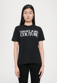 Juoda medvilninė marškinėlė su trumpomis rankovėmis, apvalia iškirpte, su ryškiai baltu užrašu "VERSACE JEANS COUTURE" priekyje.