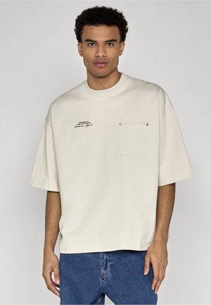 Tânăr cu păr scurt și creț poartă un tricou crem supradimensionat cu buzunar la piept și text mic negru, asortat cu blugi albaștri.