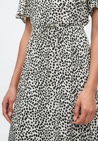 Φόρεμα με ασπρόμαυρο πουά, κοντά μανίκια, ζώνη με κορδόνι στη μέση και υφή υφάσματος που ρέει. Χέρια ακουμπισμένα στο φόρεμα.