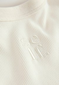 Gros plan sur un tissu côtelé blanc cassé avec des lettres brodées discrètes « se if. » près de la couture d'encolure ronde.