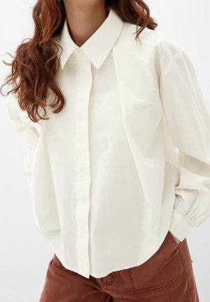 Kvinde iført en løs hvid skjorte med knapper og spidst krave samt brune bukser, poserende med den ene hånd i lommen.