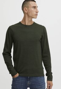 Chemise à manches longues vert foncé, col rond, tissu texturé, design ajusté, associée à un jean bleu. Aucun motif ou accent visible.