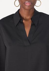 Camicia nera con colletto a V, realizzata in tessuto liscio. Presenta un colletto strutturato e una lucentezza discreta. Orecchini circolari in oro visibili.