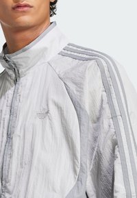 White and light gray windbreaker jacket with zip closure, high collar, textured fabric, and three vertical stripes on each shoulder.
