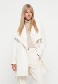 White tailored coat with a draped collar, made of a soft material, featuring long sleeves and a front opening, paired with beige undergarments.
