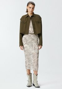 Giacca cropped verde oliva con due tasche anteriori, indossata sopra un abito beige con un sottile motivo a righe, abbinato a stivali da cowboy grigio chiaro.