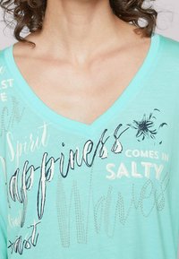 En mintgrön T-shirt med V-ringning gjord av mjukt tyg, med text och mönster i vitt, marinblått och metalliska detaljer.