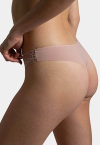 String beige à dos en dentelle réalisé dans un tissu doux et extensible, avec un devant lisse et des détails en dentelle décoratifs à l'arrière.