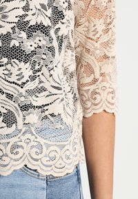 Top in pizzo beige con motivo floreale, tessuto semi-trasparente, maniche e orlo con bordi ondulati; indossato sopra jeans di denim azzurri.