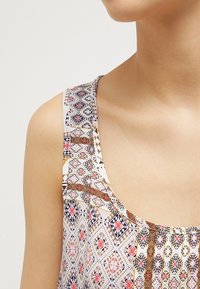 Haut sans manches à motif présentant des designs géométriques rouges, bleus et dorés sur un fond clair, avec une texture lisse et un large col.