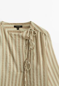 Blouse beige et blanche à rayures, manches longues, col rond et trois nœuds attachés le long de l'épaule droite devant.