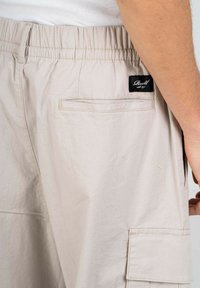 Beige Cargo-Shorts mit einem elastischen Bund, Seitentasche und einem Markenlabel auf der Rückseite, aus leichtem Stoff gefertigt.