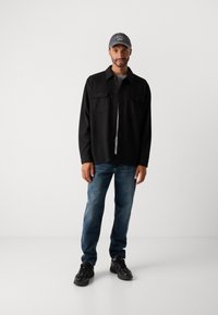 Mand iført en sort knap-op jakke, grå skjorte, blå jeans, sorte sneakers og en mørkegrå kasket, der står foran en ensfarvet baggrund.