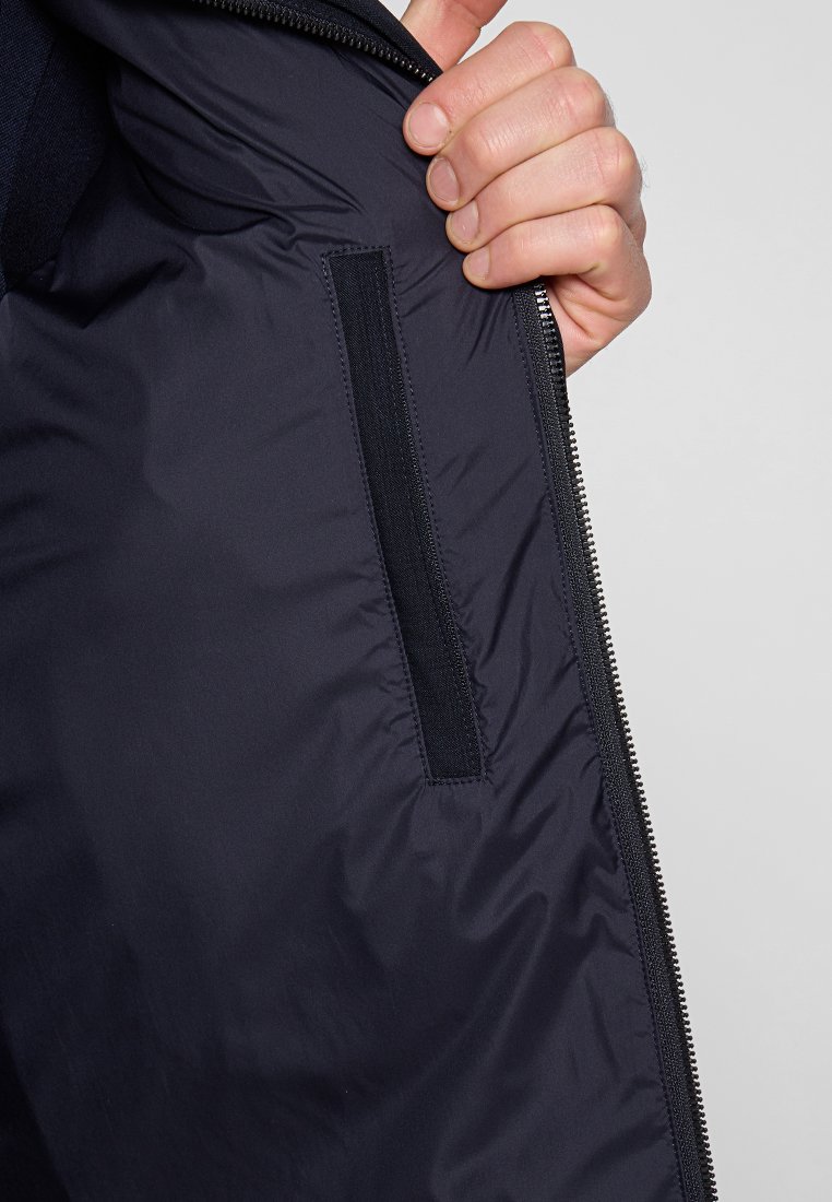 Schwarzes, isoliertes Innenfutter der Jacke mit glatter, glänzender Textur; enthält eine vertikale Tasche mit einem Stoffklappenverschluss und einem Reißverschluss entlang der Kante.