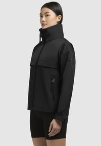 Giacca nera impermeabile con colletto alto, chiusura frontale con zip, tasca sul petto e tasche laterali con zip. Texture liscia e opaca.
