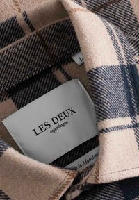 Colletto di camicia in flanella a quadri beige e blu scuro con un'etichetta bianca che mostra "Les Deux Copenhagen" e dettagli per la cura, trama del tessuto visibile.