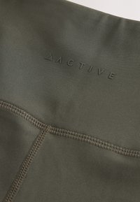 Actieve stof in olijfgroen met een gladde textuur, voorzien van een gestikte naad en een reliëf "ACTIVE" logo aan de bovenkant.