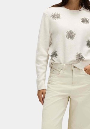 Femme portant un pull crème avec des ornements en forme d'étoile argentée, associé à un pantalon crème taille haute, main dans la poche, sur fond uni.