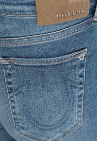 Jean en denim bleu avec une poche arrière ornée d'un motif de couture distinctif et d'une étiquette en cuir portant l'inscription "True Religion".
