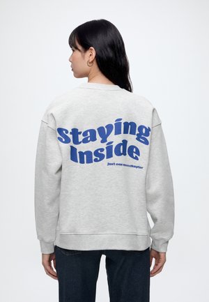 Kvinde med langt mørkt hår iført lysegrå sweatshirt med blå tekst "Staying Inside just one more chapter" på ryggen, set fra siden.