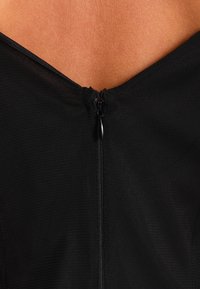 Svart åtsittande plagg med V-ringning och dragkedja i ryggen, med en texturerad, nätliknande tyg.