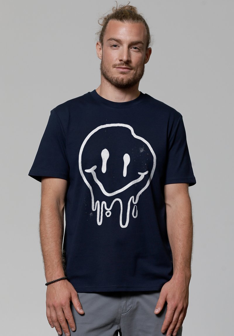 T-shirt en coton bleu marine avec un grand graphique de visage souriant éclaboussé en blanc. Manches courtes et col rond.