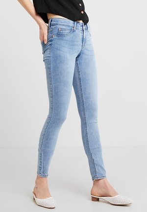 Person trägt hellblaue Skinny-Jeans und weiße, geflochtene Slip-On-Schuhe, mit einer Hand in der Hosentasche vor einem einfarbigen Hintergrund.